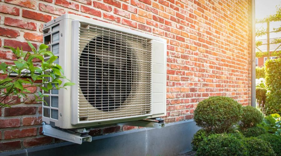 Air conditioning unit on brick wall with greenery in sunlight, showcasing modern HVAC solutions by The Bosworth Company.