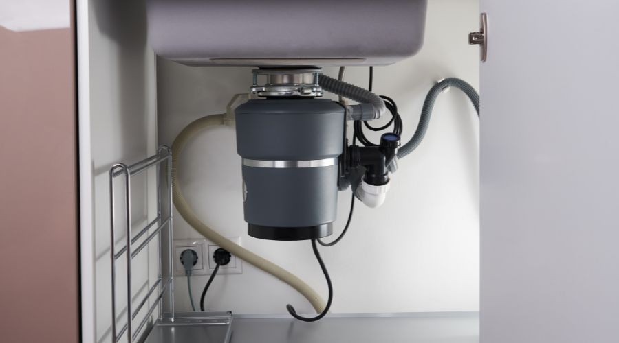 Dark gray garbage disposal under kitchen sink with visible plumbing and electrical connections, by The Bosworth Company.